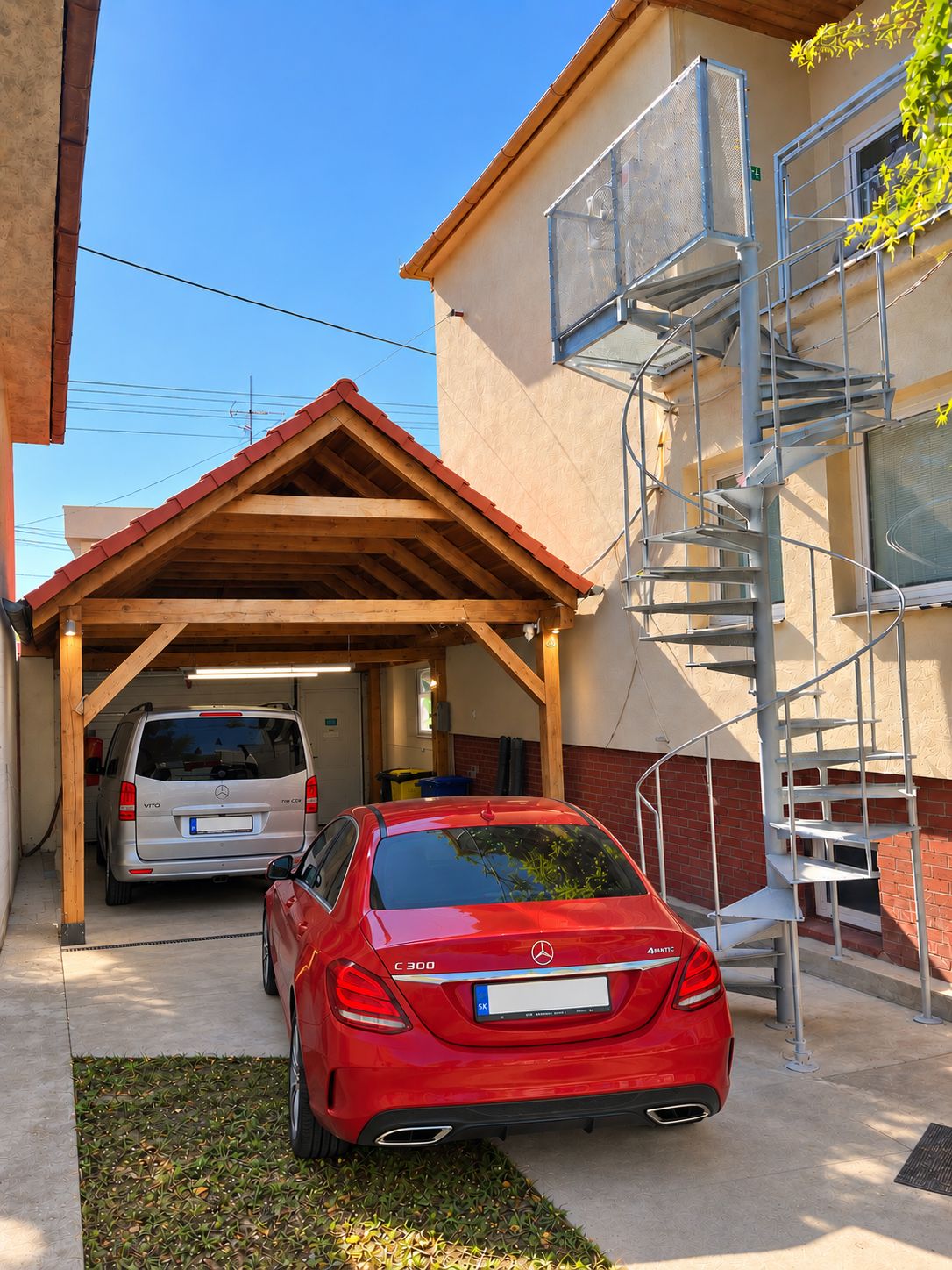 Um carro vermelho estacionado numa área coberta com teto de madeira e uma escada de metal que leva a um edifício.