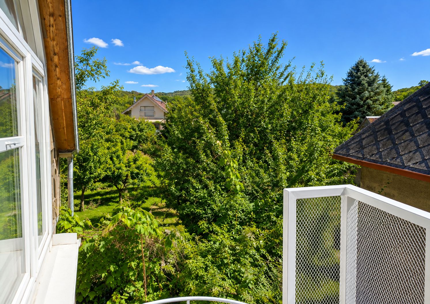 A residential property with a view of a lush green landscape, a house in the background, and a white balcony with a metal railing.