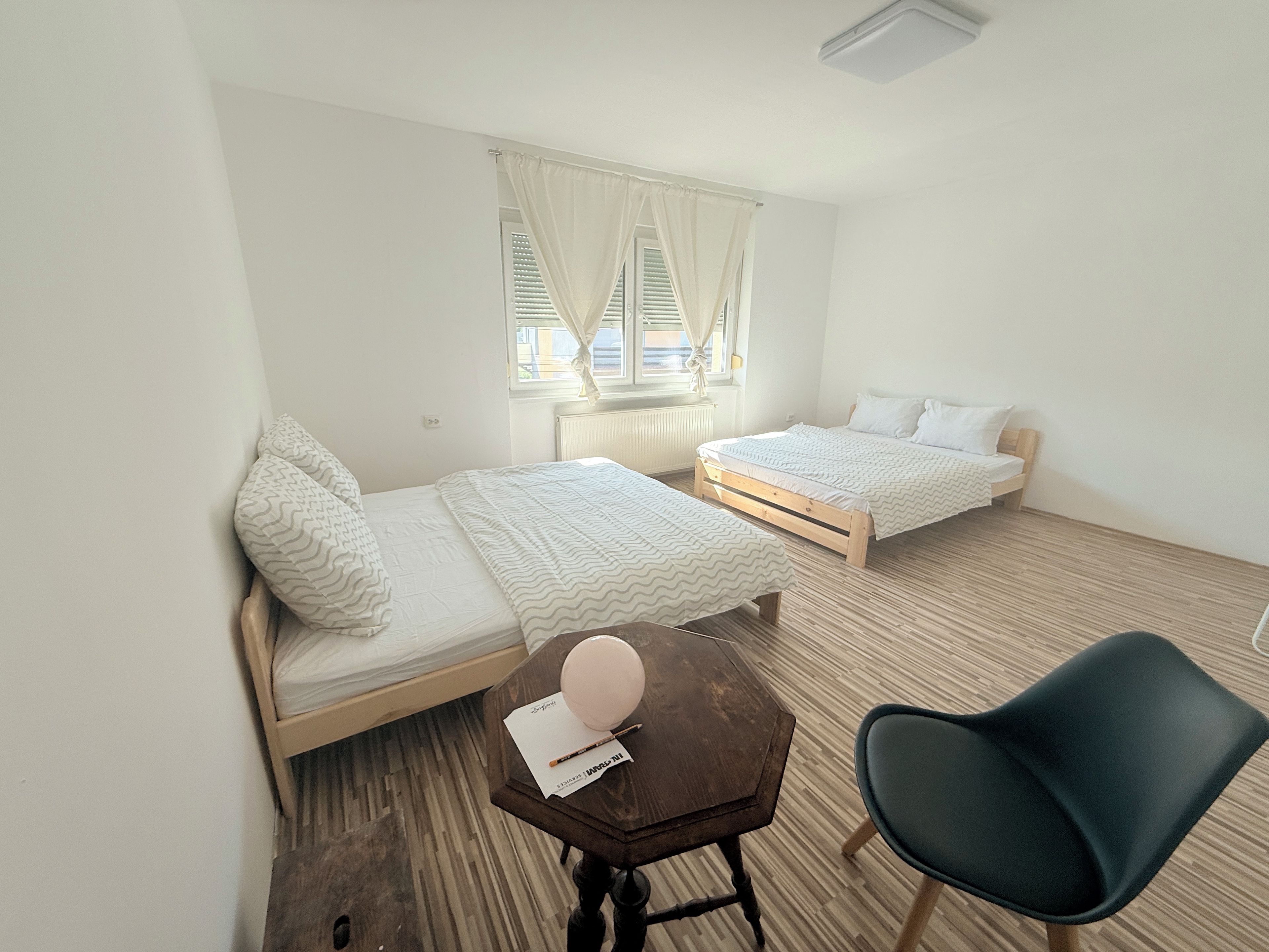 Ein Zimmer mit zwei Einzelbetten, einem kleinen Tisch mit Stuhl und einem Fenster mit Vorhängen.