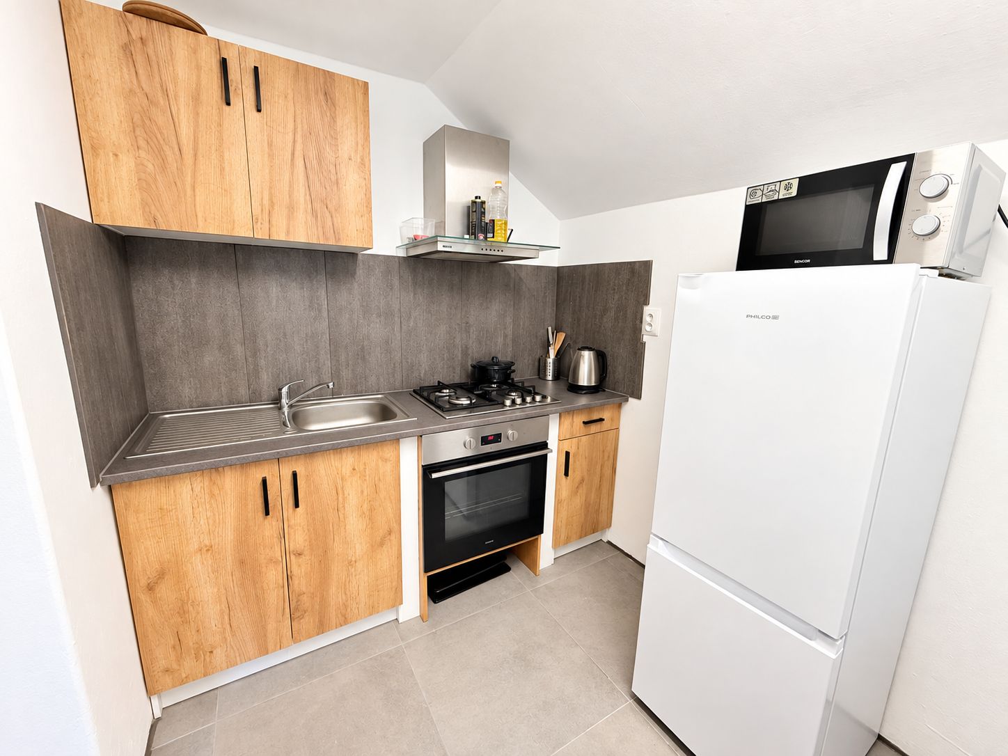 A compact kitchen with wooden cabinets, a stainless steel sink, a gas stove, a microwave, and a white refrigerator.