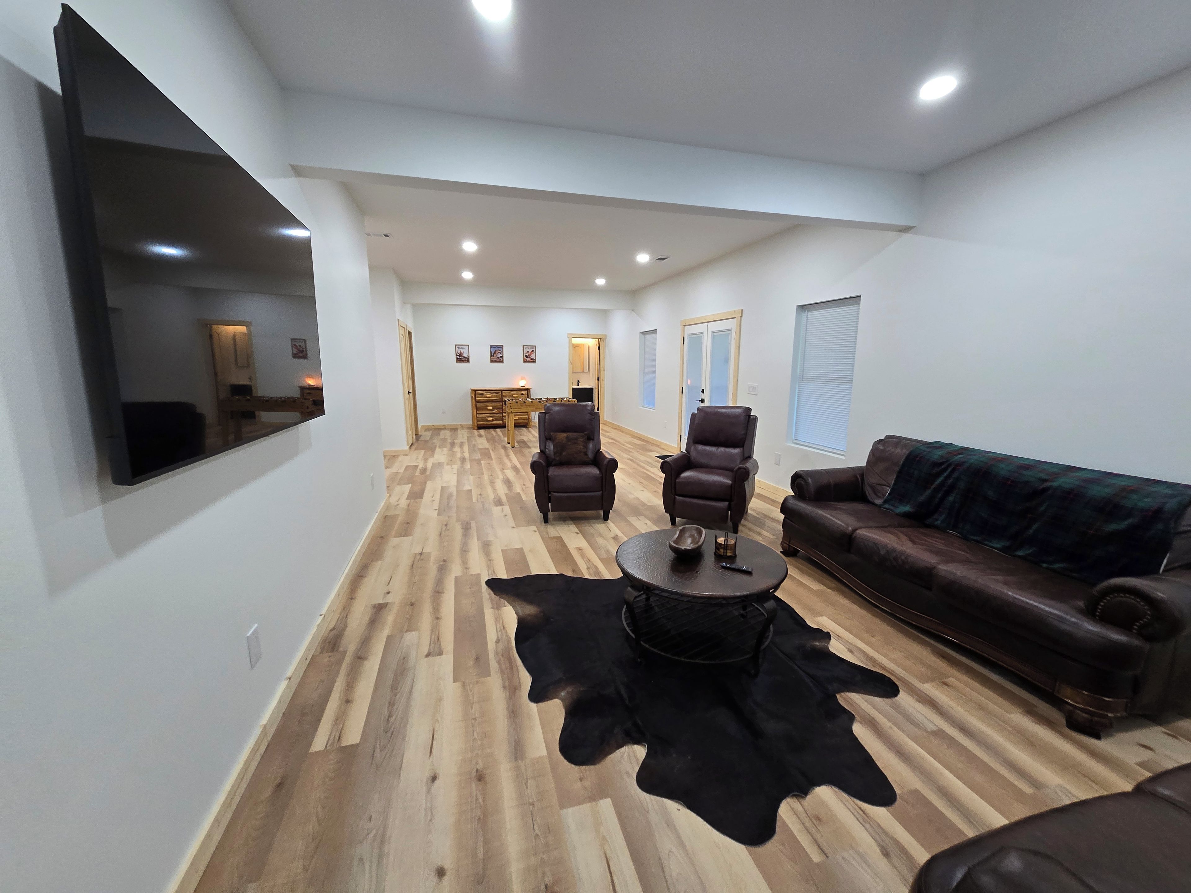 A spacious living room with a large flat-screen TV mounted on the wall, a leather sofa, two armchairs, a coffee table, and a wooden floor.