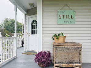 Un porche avec une enseigne indiquant « The STILL House », un panier en osier rempli de plantes et une chaise en osier.