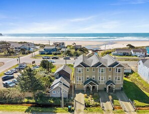A residential area with a large two-story house in the foreground, a beach in the background, and a road with parked cars. - Ocean-View Coastal Retreat | Walk to Beach | Garage + EV Charging (Rockaway Beach)