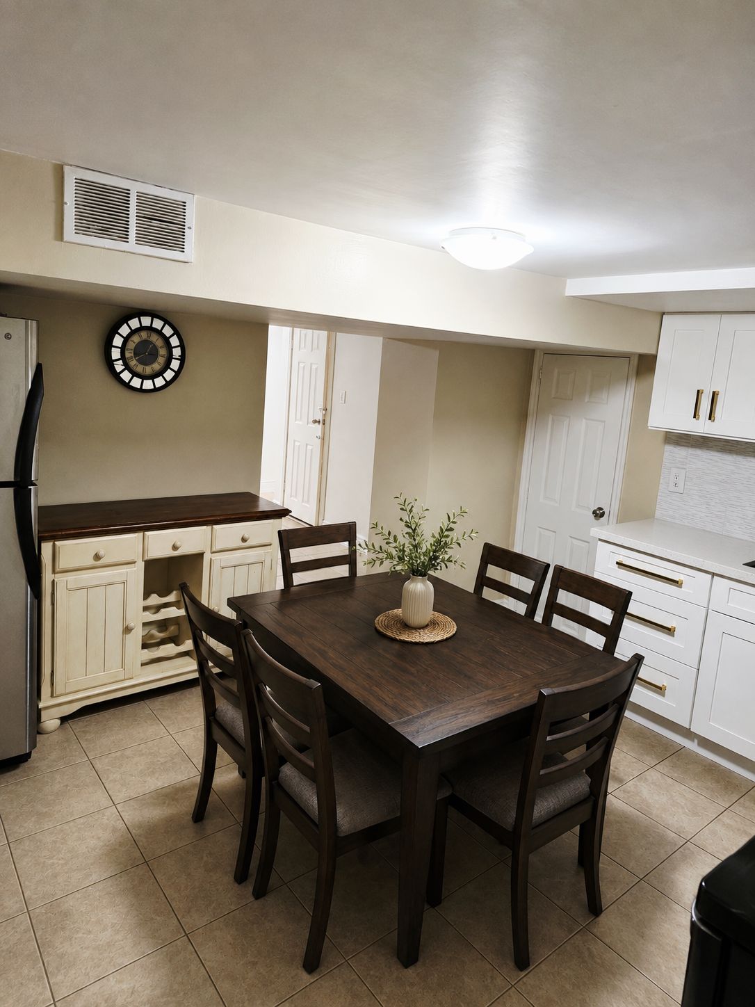 A kitchen with a dining table, chairs, cabinets, a clock, and a refrigerator.
