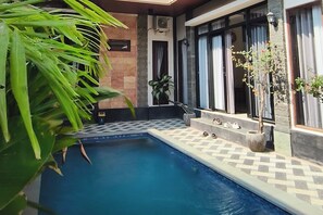 Una piscina con borde de baldosas, una planta en maceta y una ventana con cortinas.