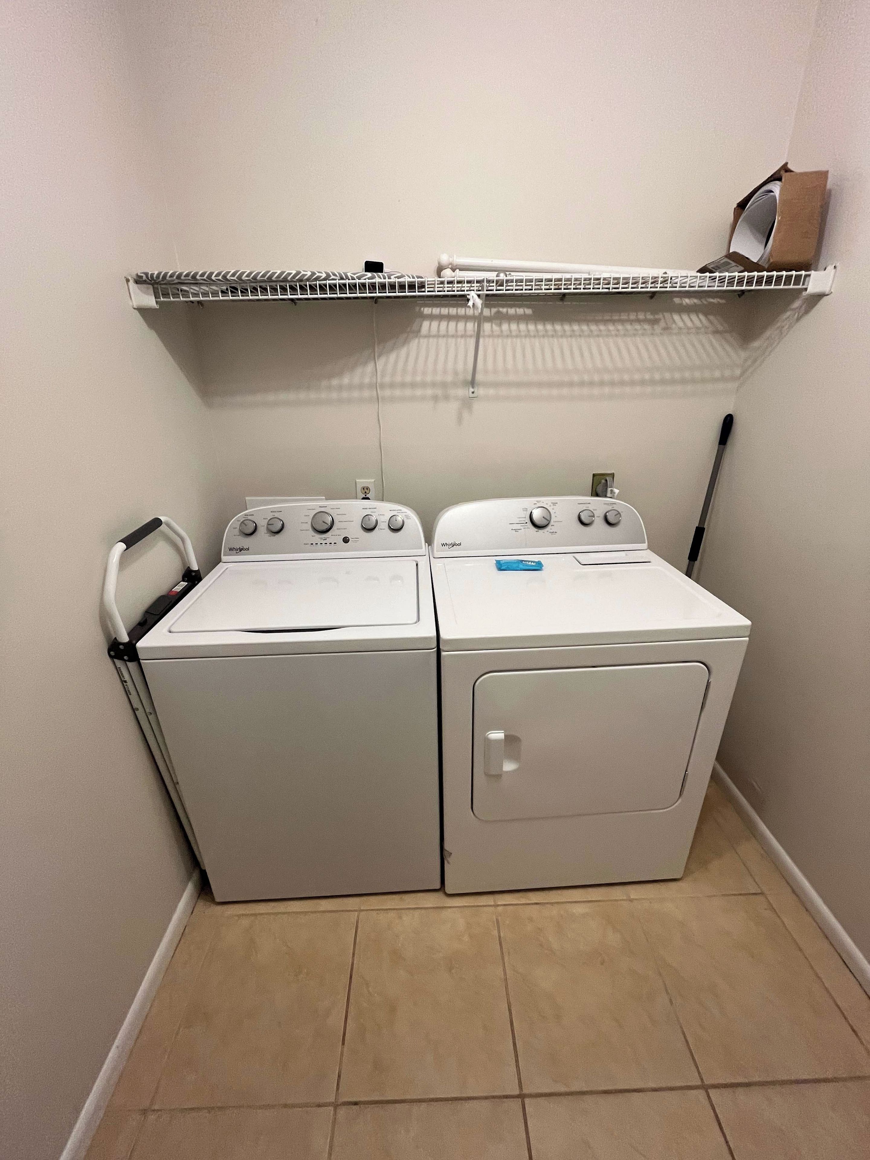 Une buanderie équipée de deux machines à laver et de deux sèche-linge à chargement frontal, superposés, avec une étagère en métal au-dessus et un balai appuyé contre le mur.