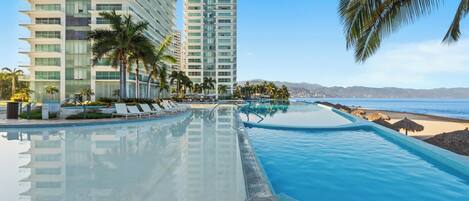 A swimming pool with lounge chairs and palm trees, adjacent to a high-rise building and a beach.