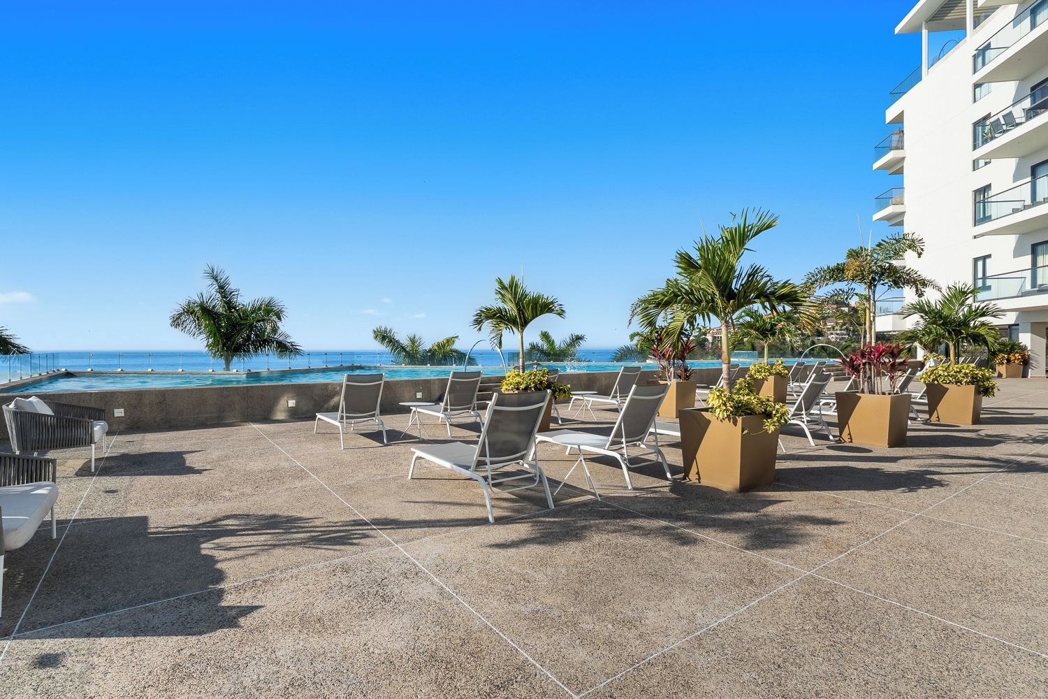 A rooftop terrace with lounge chairs and potted plants, overlooking a pool and ocean view.