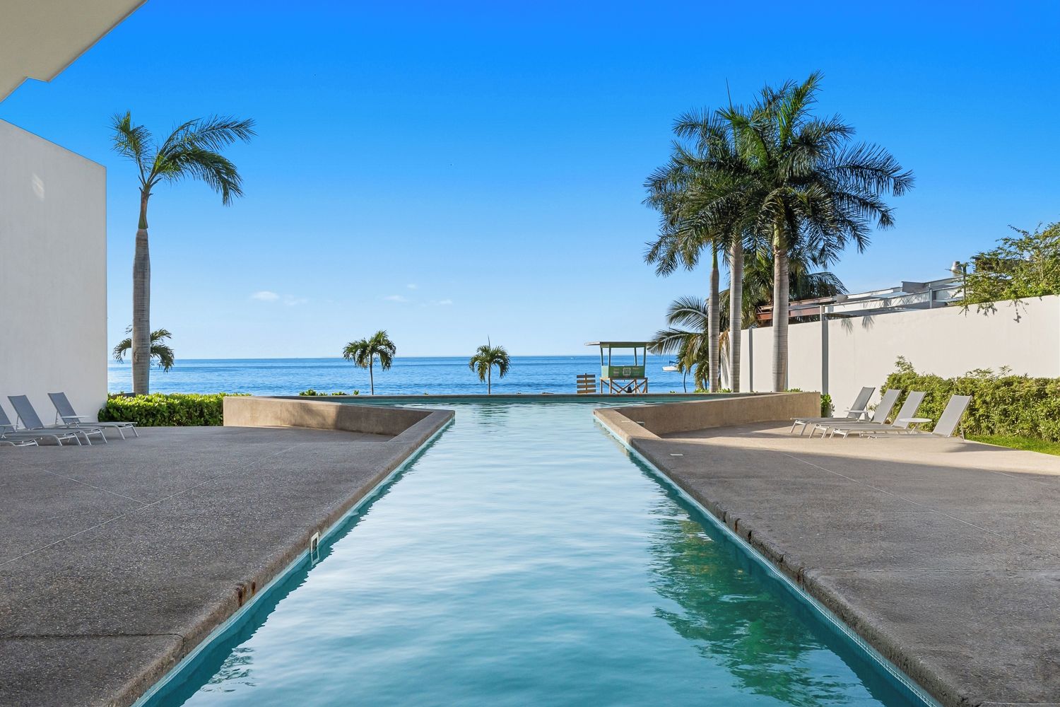 A swimming pool with a view of the ocean, surrounded by palm trees and lounge chairs.