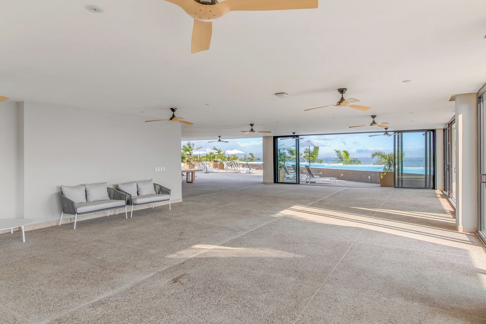 Un espace intérieur spacieux, doté d’un ventilateur de plafond, d’un canapé et offrant une vue sur une piscine et l’océan.