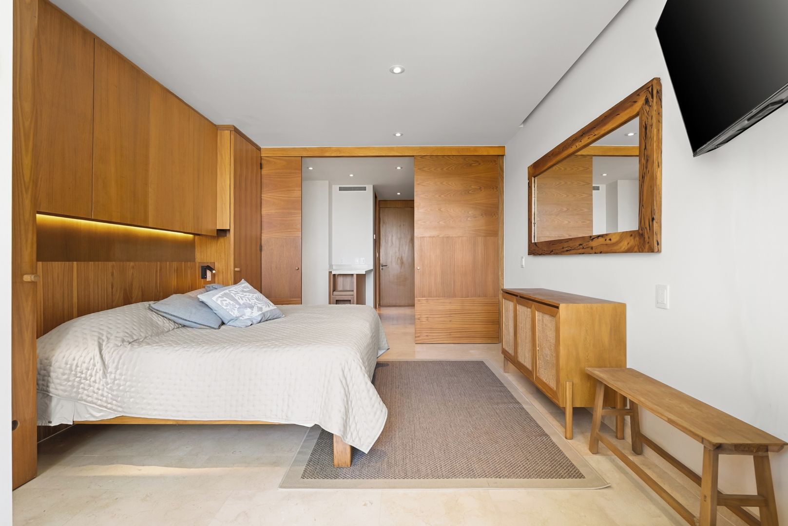 A bedroom with a bed, a wooden wardrobe, a desk, a bench, and a television mounted on the wall.