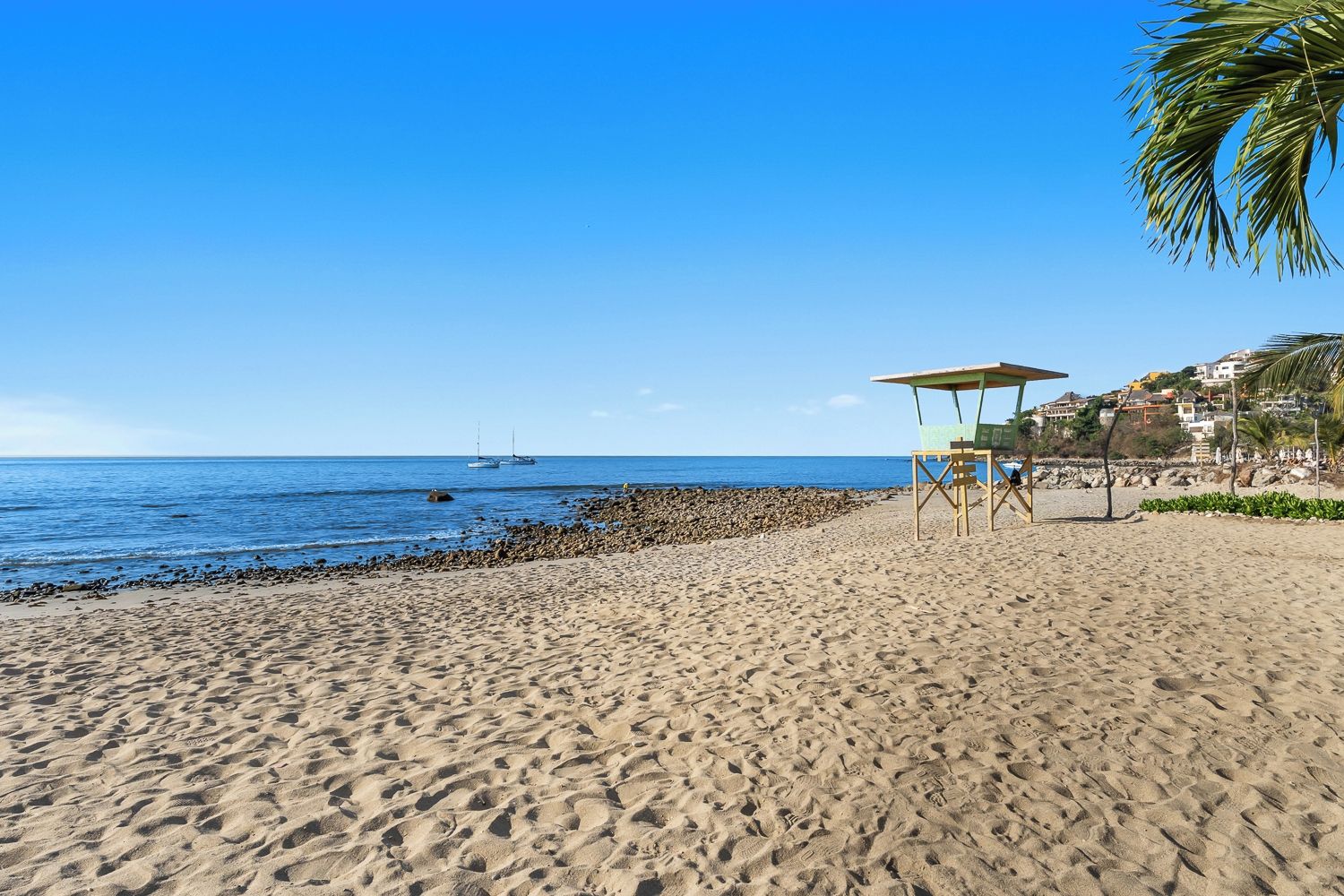 Een zandstrand met een helderblauwe lucht, een rotsachtige kustlijn en enkele boten in de verte.