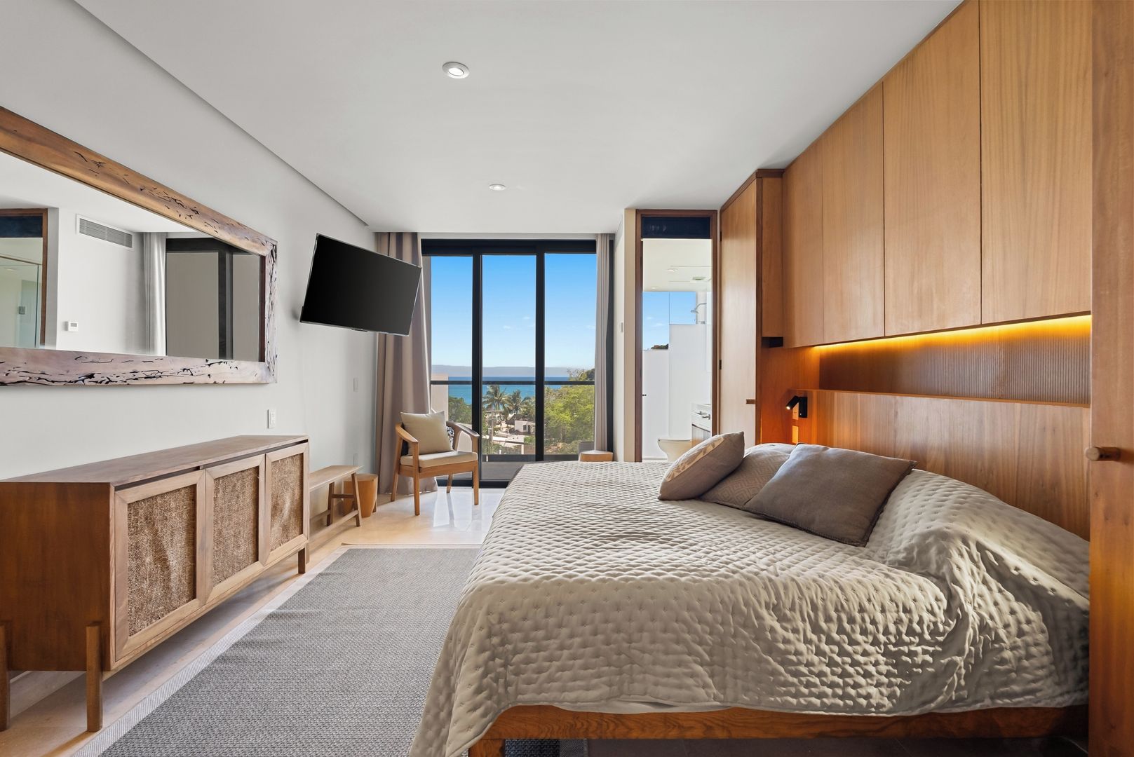 A modern bedroom with a large bed, a flat-screen TV, a wooden cabinet, and a balcony with a view.
