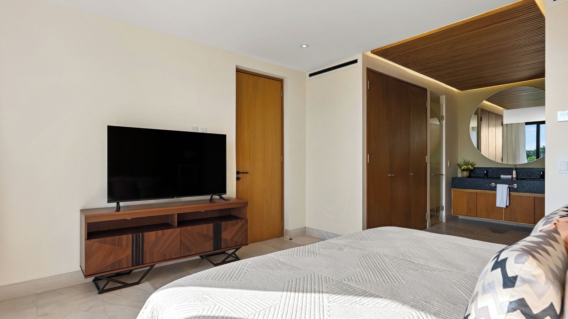 A modern bedroom with a large bed, a flat-screen TV on a wooden stand, a walk-in closet, and a bathroom with a mirror and sink.