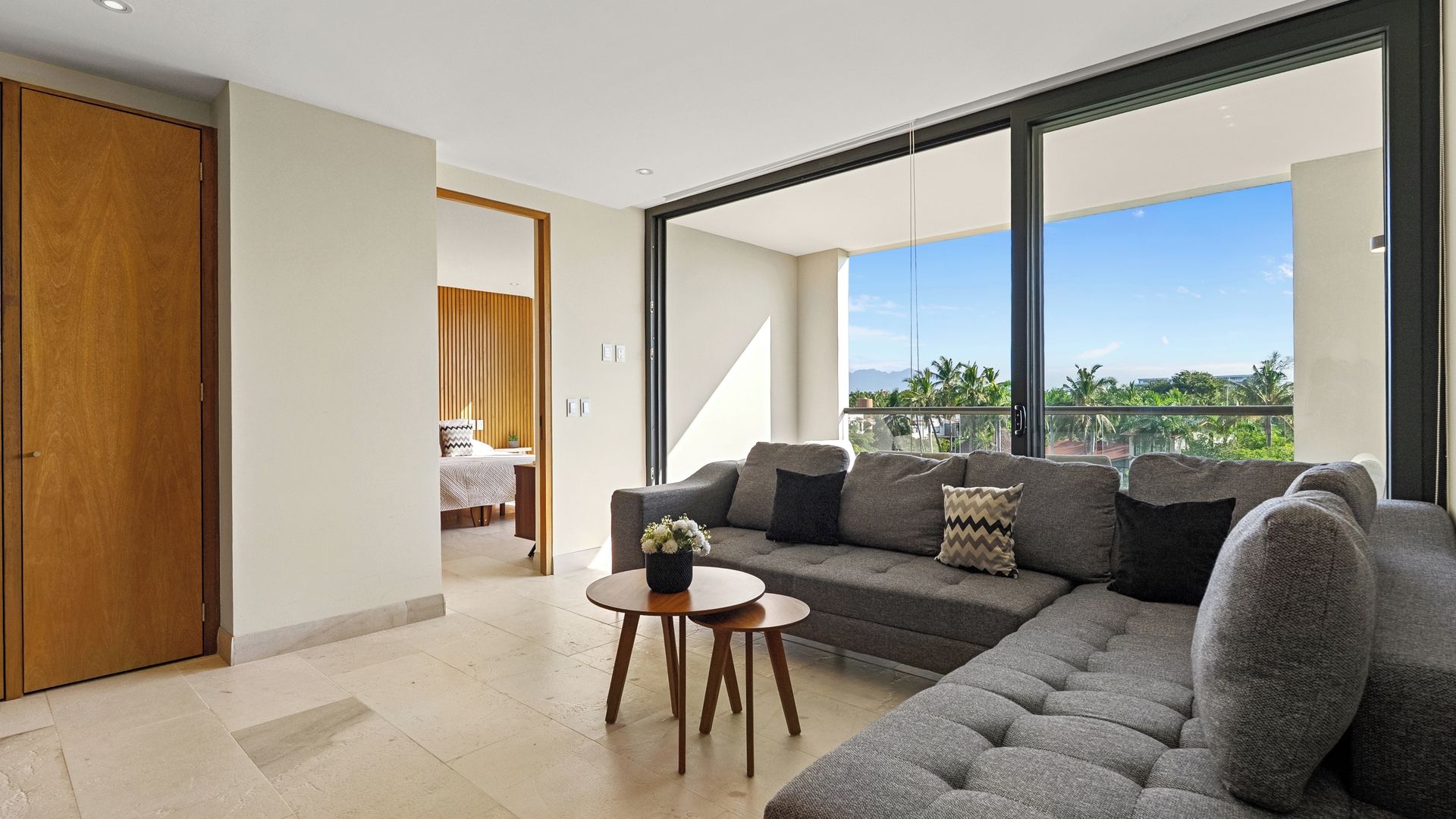 A modern living room with a grey sectional sofa, a small round table with a vase, and a view of greenery and mountains through large windows.