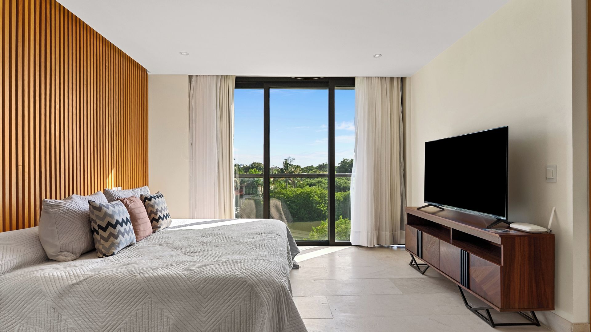 A modern bedroom with a large bed, a flat-screen TV, and a balcony with a city view.