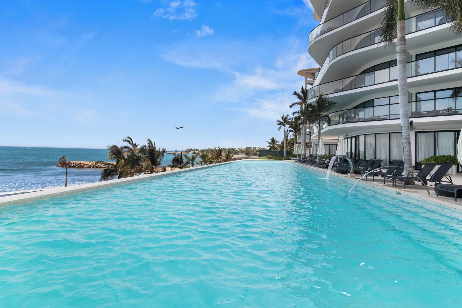 A clear blue pool with lounge chairs and palm trees, overlooking a beach and ocean.