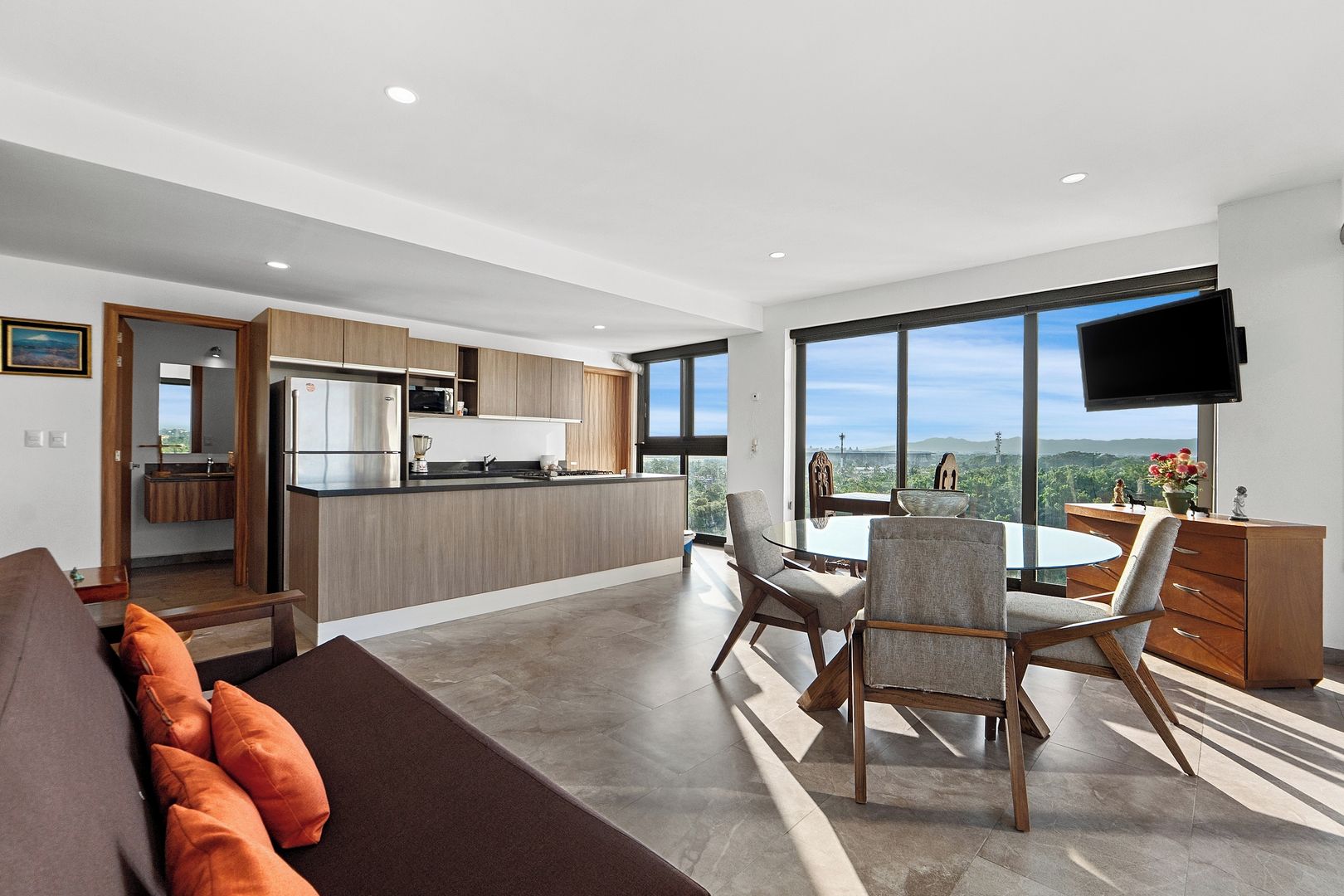 A modern kitchen with a dining area, large windows, and a flat-screen TV.