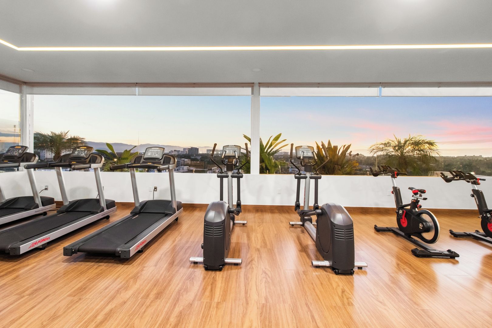 A gym with treadmills, elliptical machines, and stationary bikes, featuring a city view through large windows.
