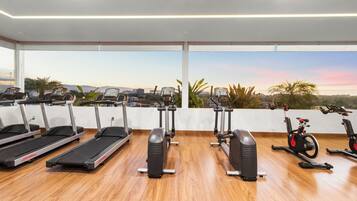 A gym with treadmills, elliptical machines, and stationary bikes, featuring a city view through large windows.