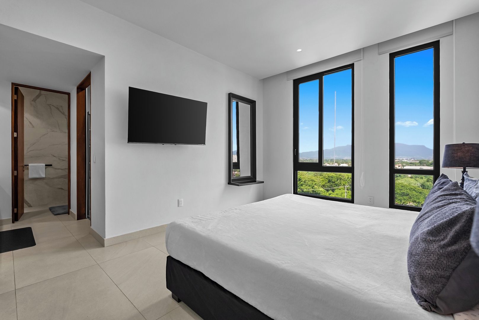 A modern bedroom with a large bed, a flat-screen TV, a mirror, and a view of greenery through large windows.