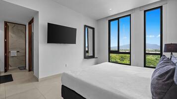 A modern bedroom with a large bed, a flat-screen TV, a mirror, and a view of greenery through large windows.