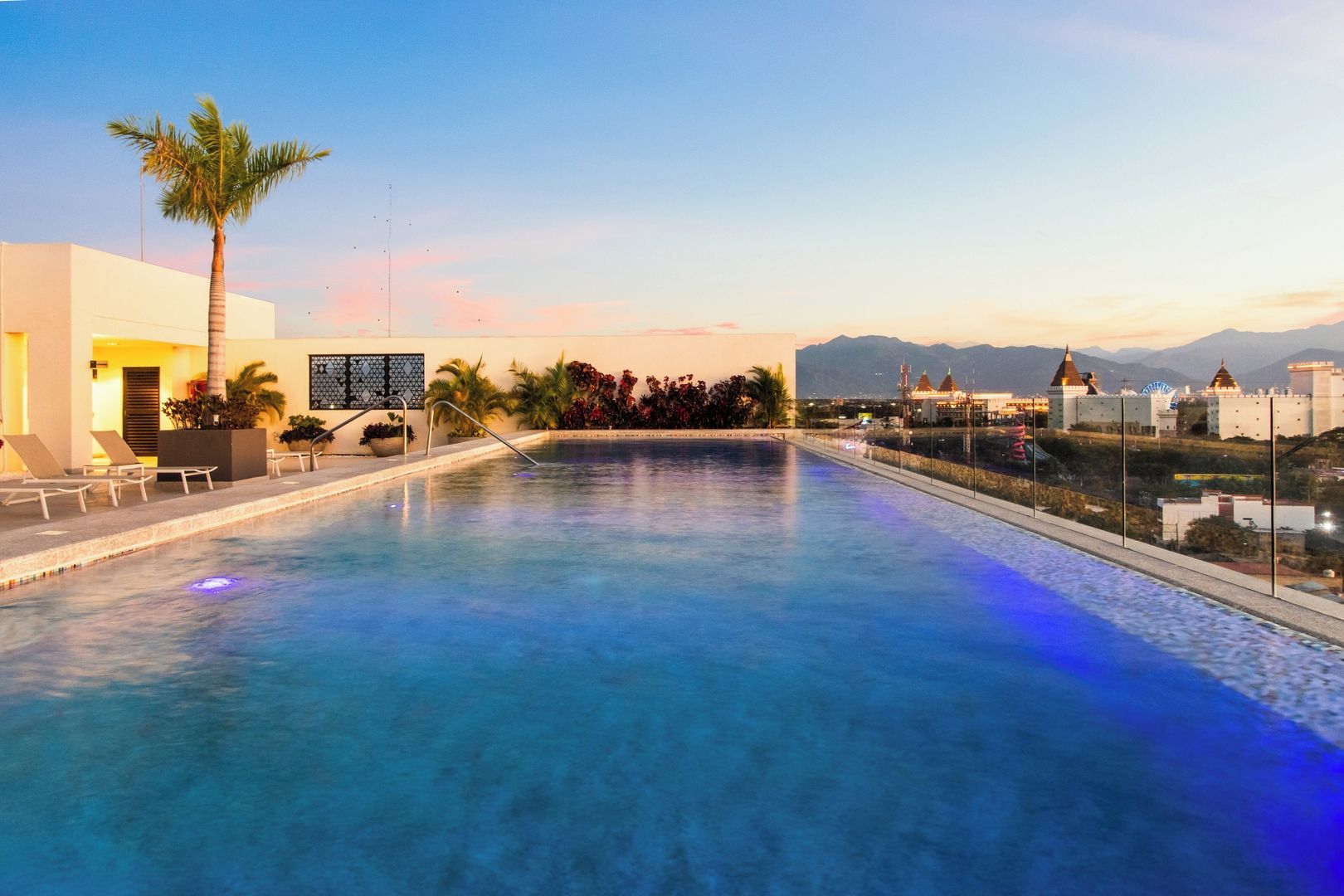 A swimming pool with lounge chairs, palm trees, and a view of mountains and buildings in the background.