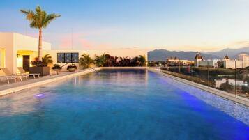 A swimming pool with lounge chairs, palm trees, and a view of mountains and buildings in the background.