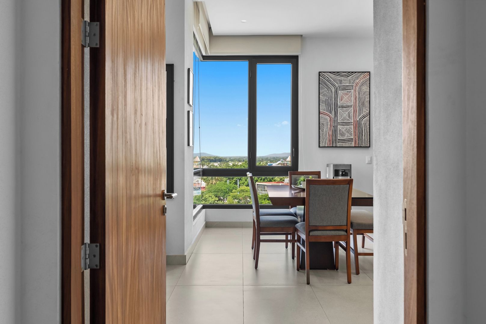 A balcony with a dining table and chairs, a view of greenery and buildings, and a modern interior with a framed artwork.
