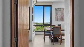 A balcony with a dining table and chairs, a view of greenery and buildings, and a modern interior with a framed artwork.