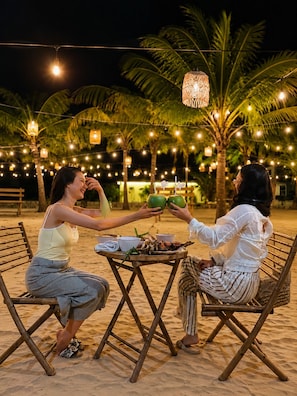 Two people seated at a table with a tropical setting, including palm trees and string lights.