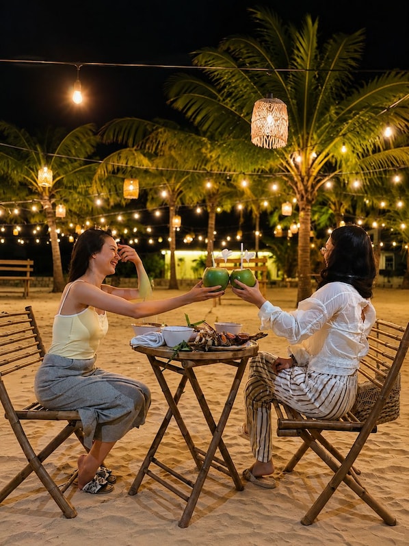 Two people seated at a table with a tropical setting, including palm trees and string lights.