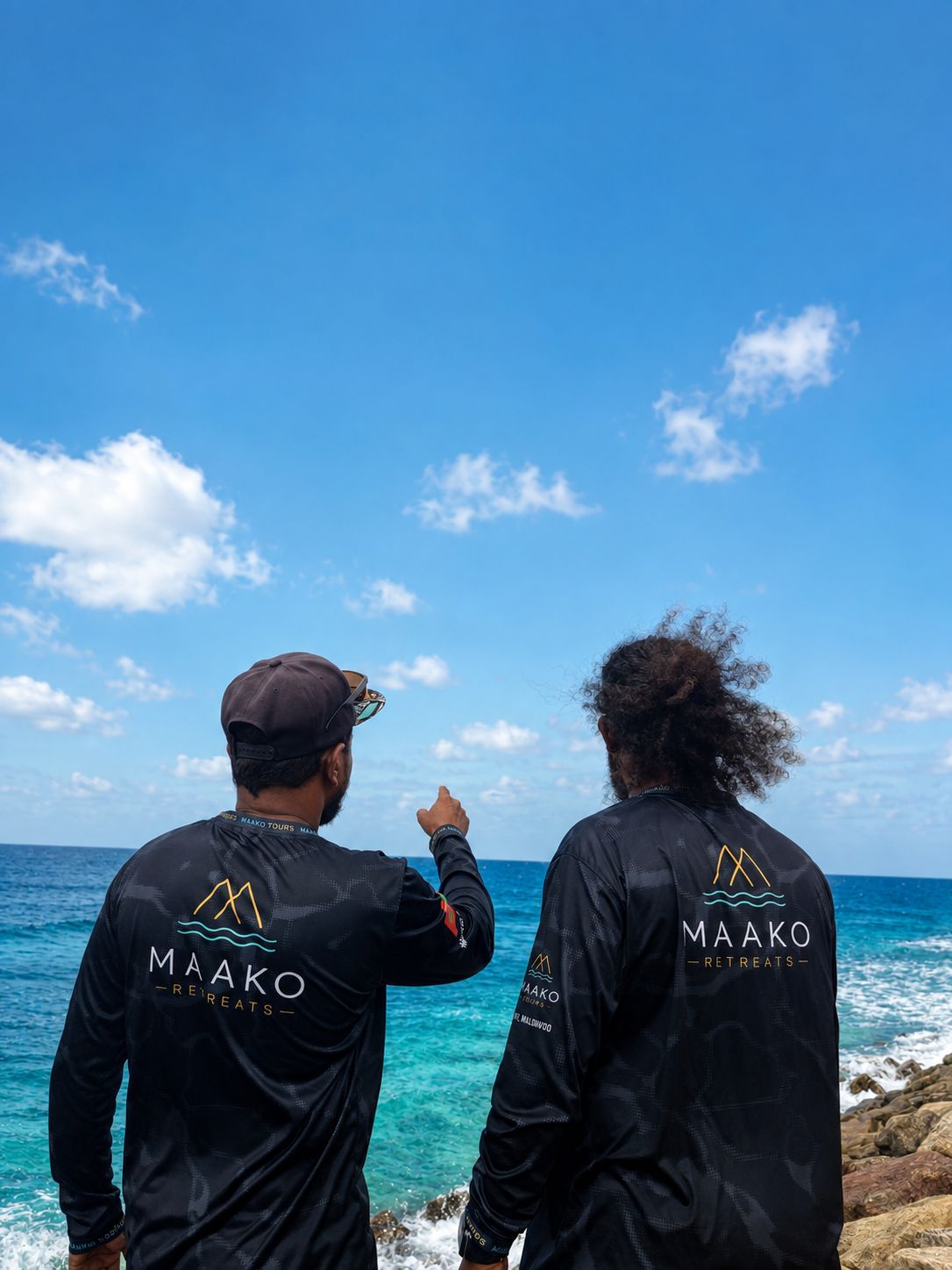 Two individuals wearing black Maako Retreats branded long-sleeve shirts, standing by the ocean with a clear sky.