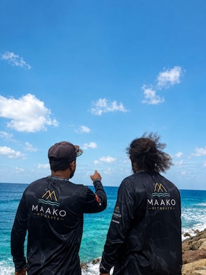 Two individuals wearing black Maako Retreats branded long-sleeve shirts, standing by the ocean with a clear sky.