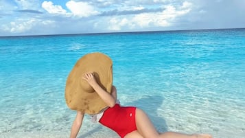 A person sitting on a beach with a large hat on their head, wearing a red swimsuit, and the ocean and sky in the background.