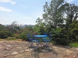 A stone patio with blue metal chairs and a table, surrounded by lush greenery and trees. - Sleeps5-PetFriendly-CoalFire-Snowdonia (Dolgellau)