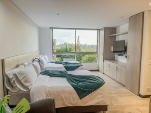 A modern hotel room with two beds, a television, and a large window offering a view of greenery. - Llanogrande 1BR Suite Near MDE Airport AC (Rionegro)