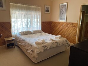 A hotel room with a bed, a small bedside table, a window with sheer curtains, and wooden paneling on the walls.