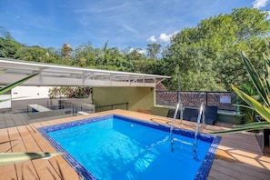 Ein Schwimmbecken mit Holzterrasse, einer überdachten Sitzgelegenheit und Blick auf üppiges Grün.