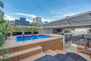 Ein Pool auf dem Dach mit Holzterrasse, einer Bar mit Sitzgelegenheiten und Blick auf die umliegenden Gebäude.