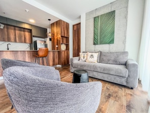 Ein modernes Wohnzimmer mit einem grauen Sofa, zwei grauen Sesseln und einem Couchtisch. Es gibt eine Küchenzeile mit Holzschränken und einer Pendelleuchte.