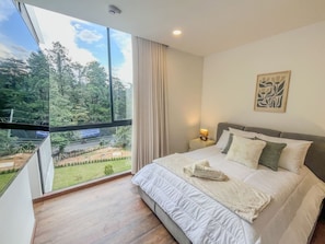 Ein Schlafzimmer mit großem Fenster, einem Bett mit weißer Bettwäsche, einem Nachttisch mit Lampe und einem gerahmten Kunstwerk an der Wand.