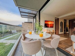 Ein moderner Essbereich im Freien mit einem runden Tisch, gepolsterten Stühlen und einem Glasgeländer mit Blick auf einen Garten.