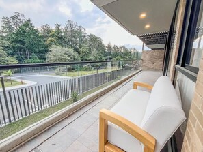 A balcony with a view of a road and trees, a glass railing, and two white cushioned chairs. - El Retiro Design Forest Panoramic View (Retiro)