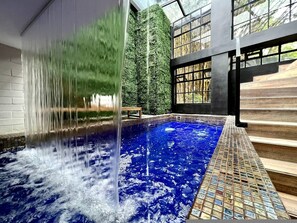 Ein Innenpool mit Wasserfall, umgeben von moderner Architektur mit großen Fenstern und Blick auf Bäume.