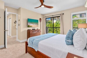 Ein Schlafzimmer mit einem Bett, einem Deckenventilator, einem Gemälde, einer Holzkommode und Blick auf Grün durch ein Fenster.