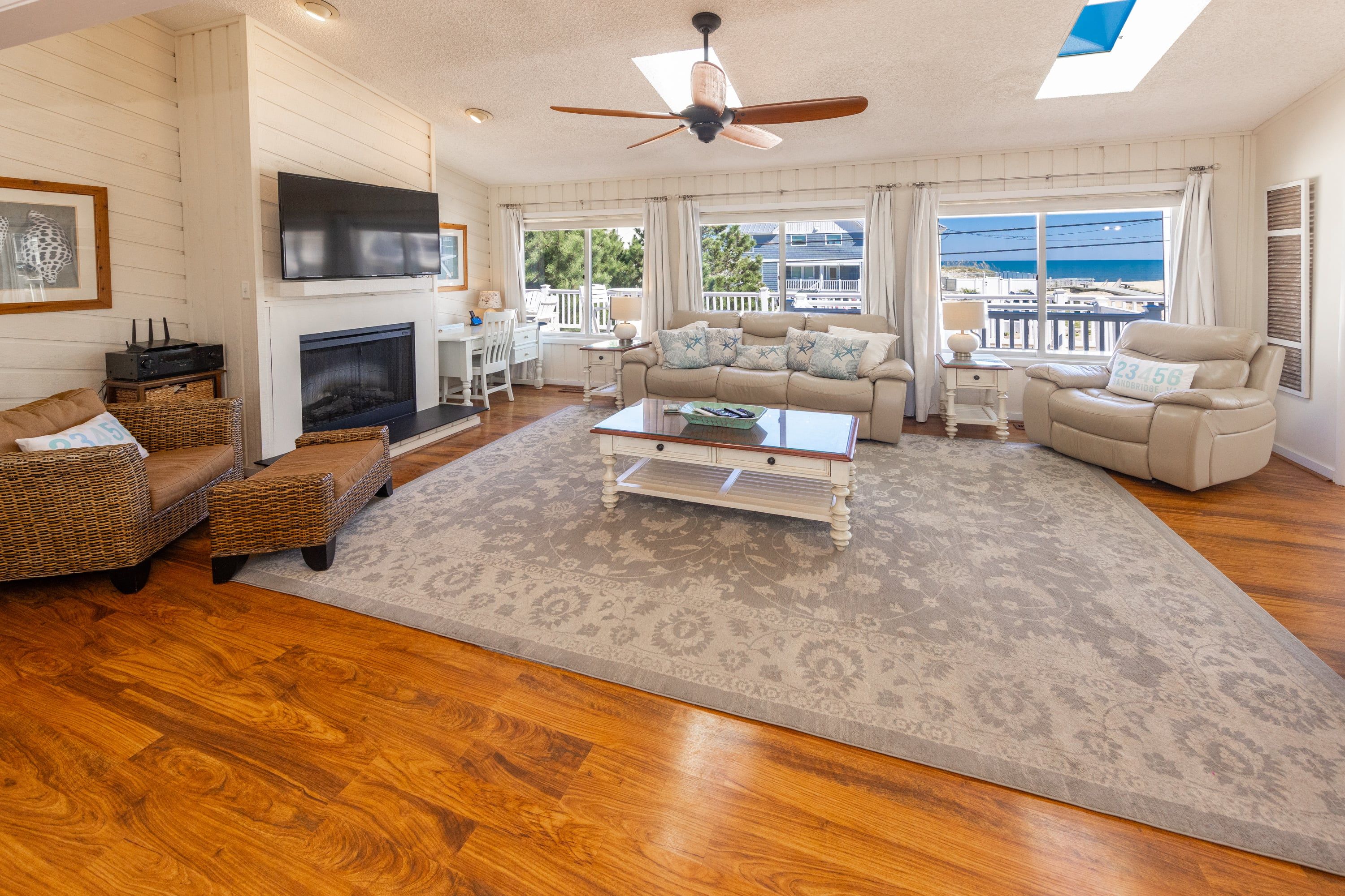 A spacious living room with a fireplace, a ceiling fan, and large windows offering a view of the sea.