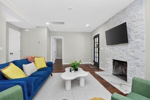 A living room with a blue sofa, a white coffee table, a fireplace, and a flat-screen TV mounted on a stone wall.