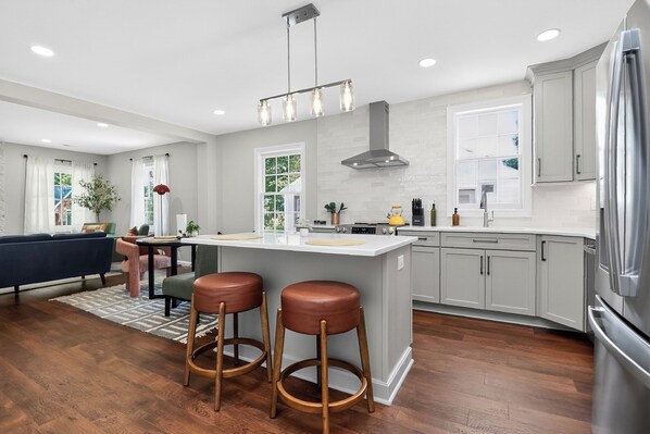 A modern kitchen with a central island, bar stools, a dining area with a sofa, and a large window providing natural light.