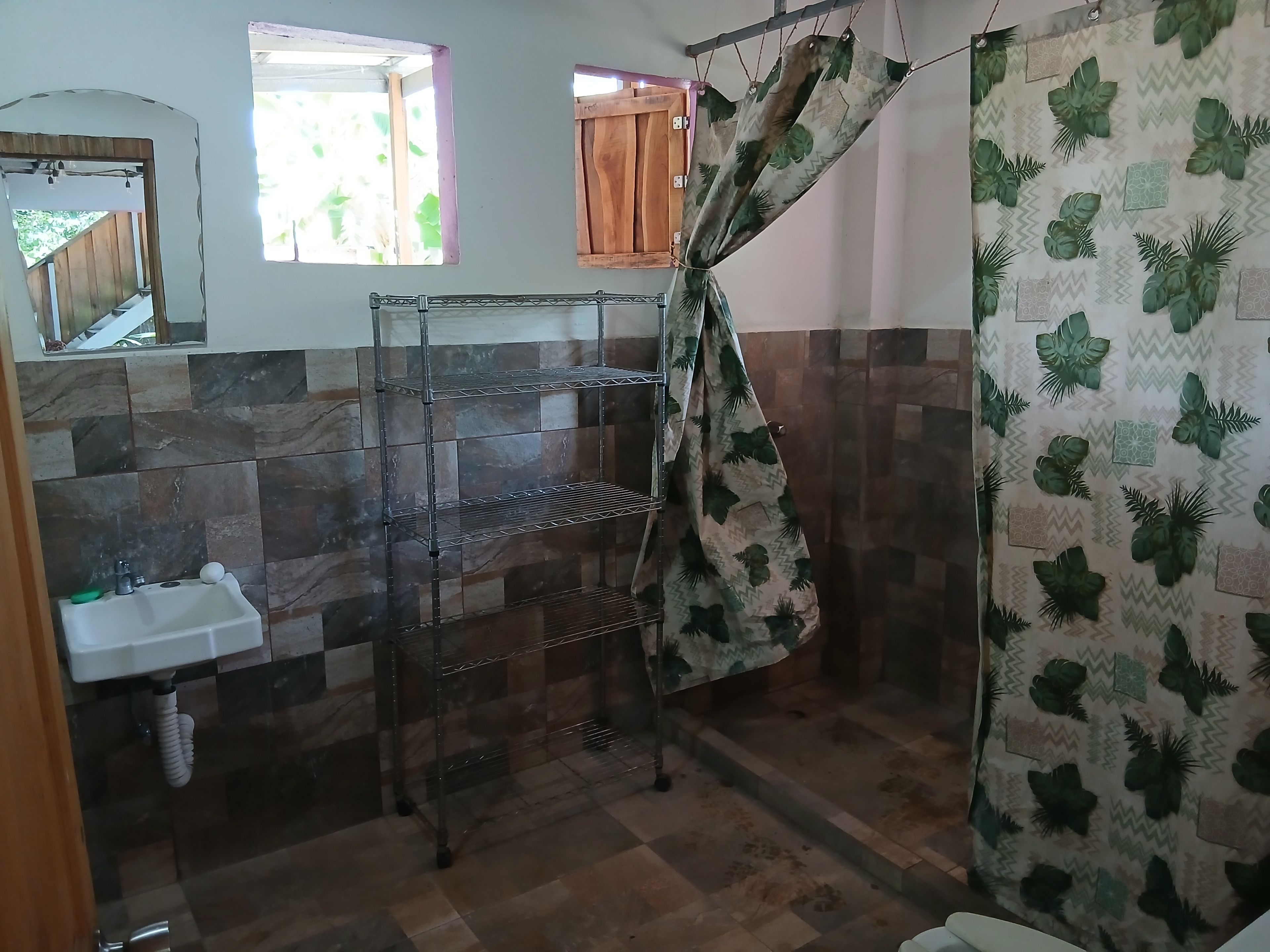 Une salle de bain avec un sol carrelé, un lavabo, une étagère en métal et un rideau de douche orné d’un motif de feuilles.